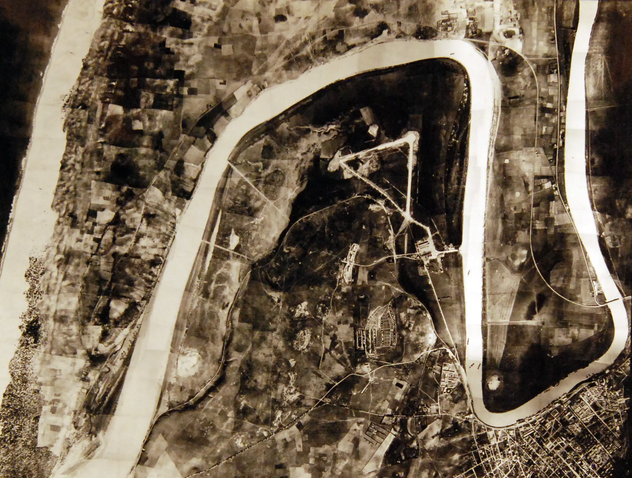 An aerial black and white photograph, showing a snaking river surrounded by rural roads with a city grid in the bottom right.