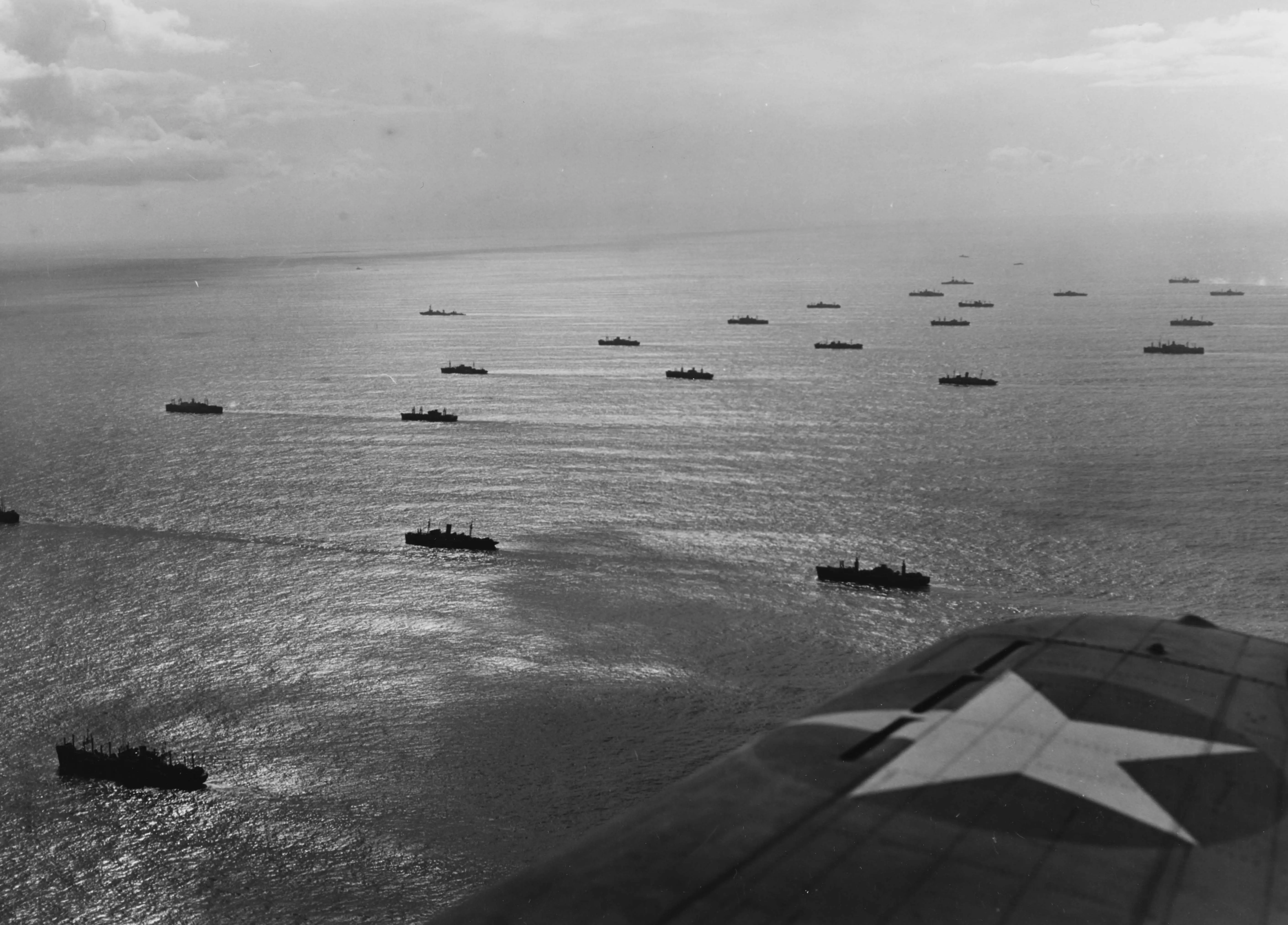 An aerial photograph of about two dozen ships steaming in open ocean and lined up in several columns. The wing of the plane is visible with the circle and star roundel of US war planes.