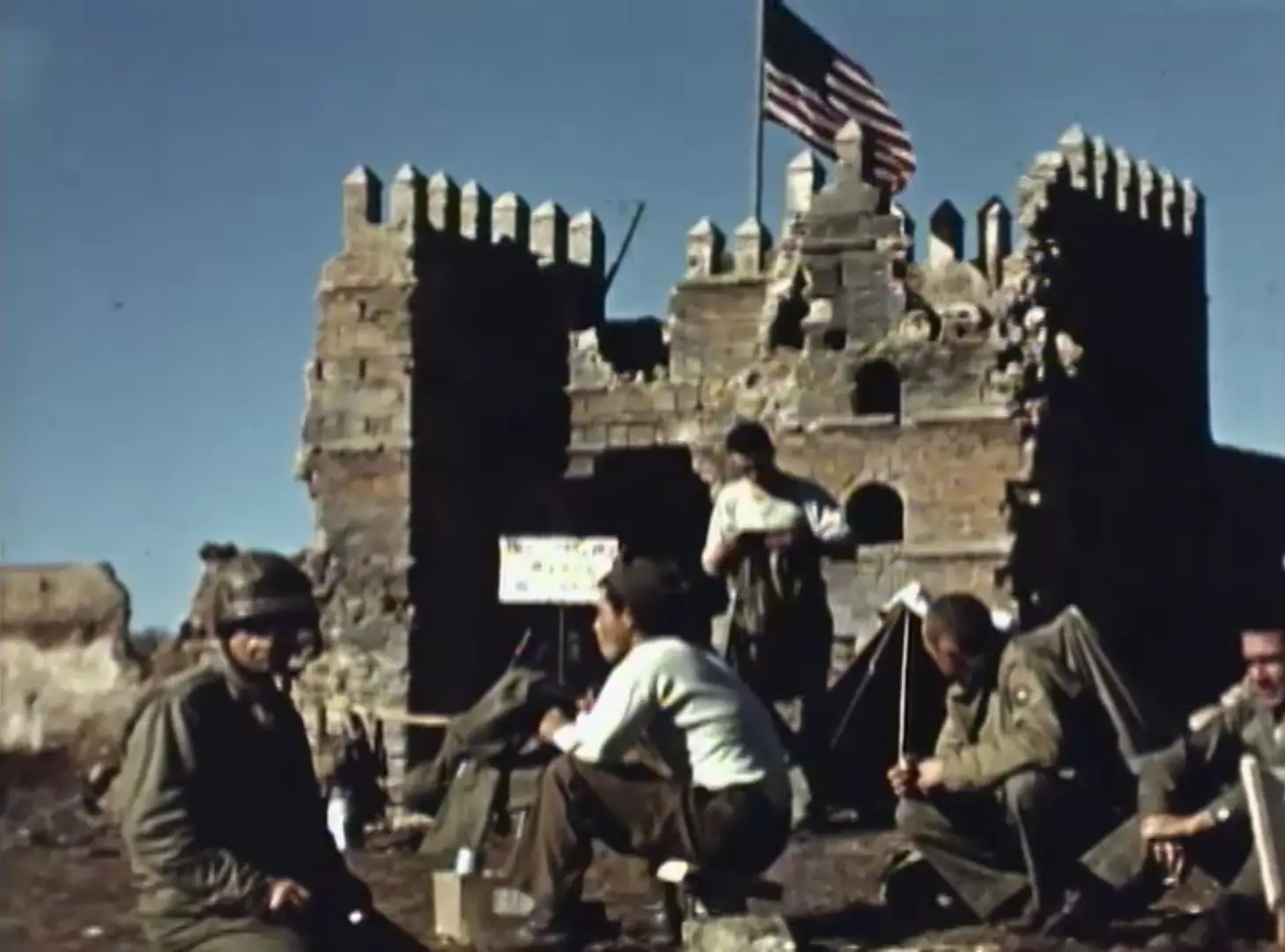 A fuzzy still taken from color film showing American soldiers resting outside a severely damaged stone gatehouse with an American flag flying above.