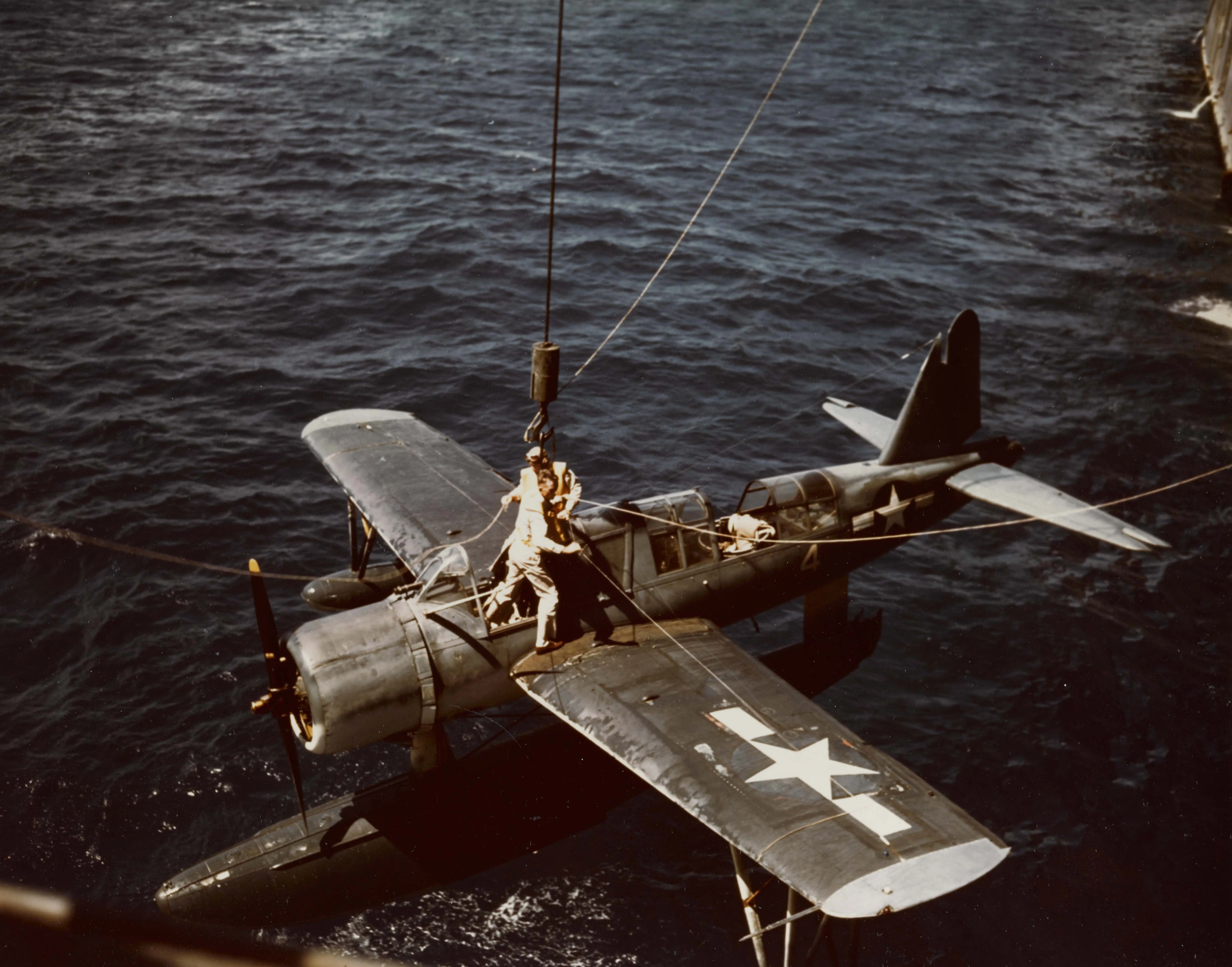 A color photograph of a float plane being lifted out of the water by a crane. The pilot and copilot have climbed out of the plane and are securing the plane to the crane hook.