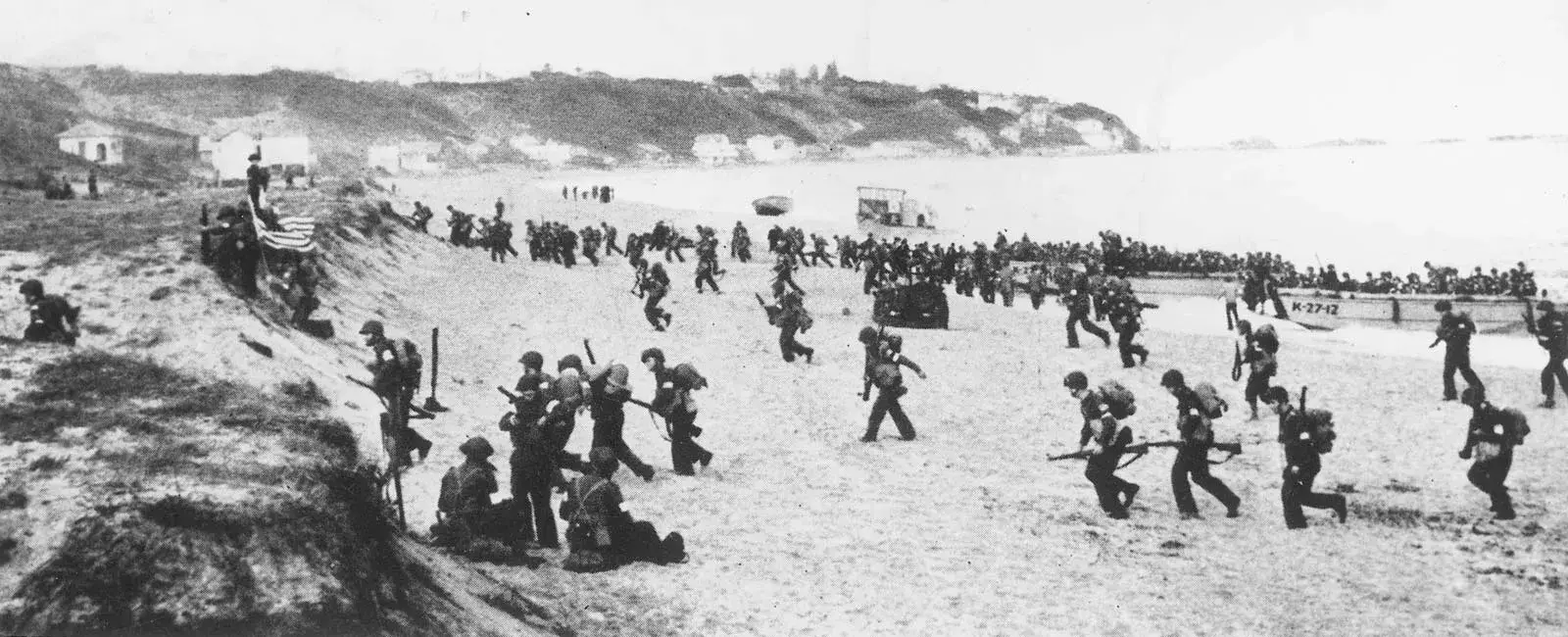 A black and white photograph of American soldiers landing on a beach. Landing craft are visible on the right and still unloading, other soldiers are approaching a small slope. Loosely scattered houses are visible in the distance.