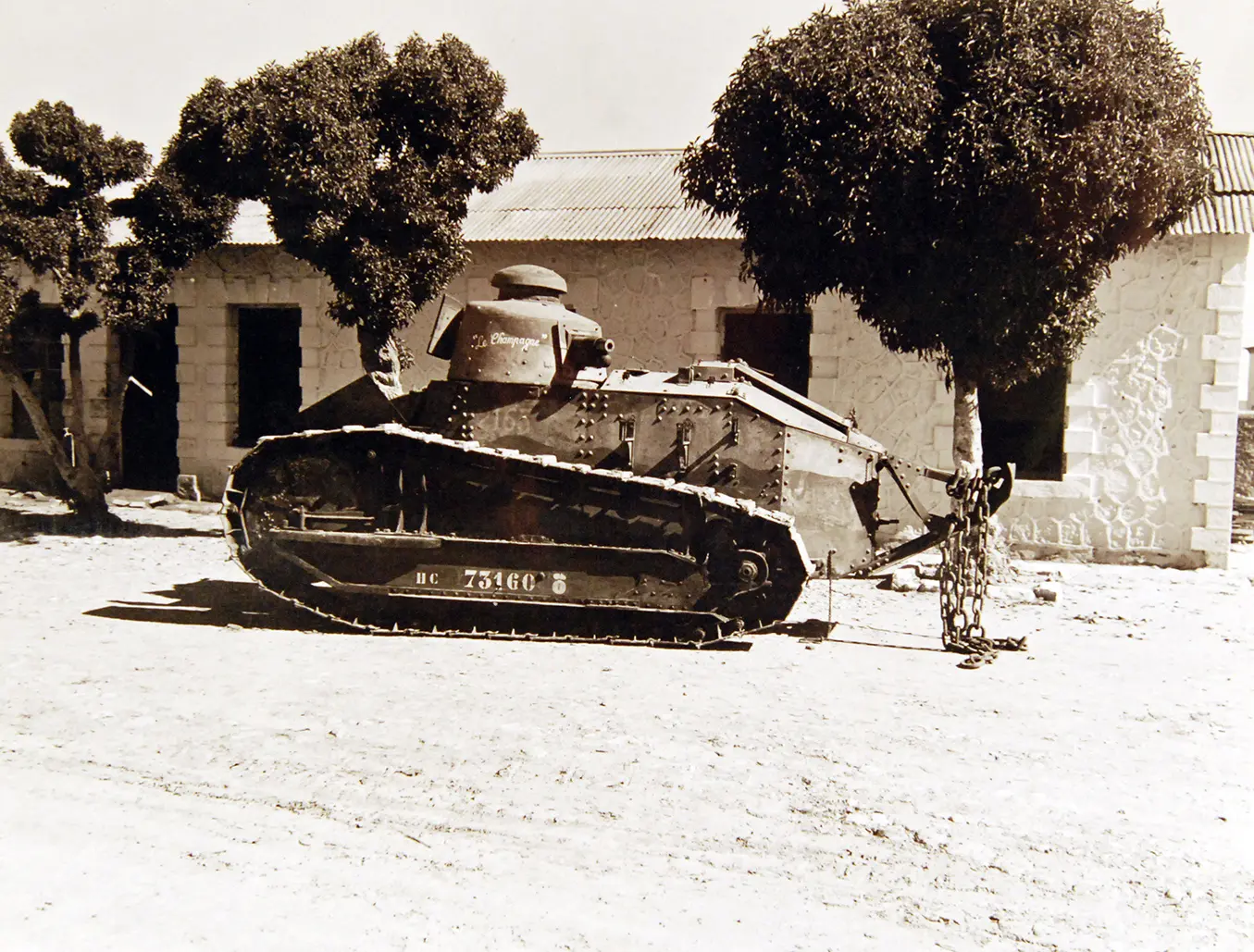 A black and white photograph of a French Renault tank, parked in front of a stone building. The tank has the words Le Champagne artfully written on its turret