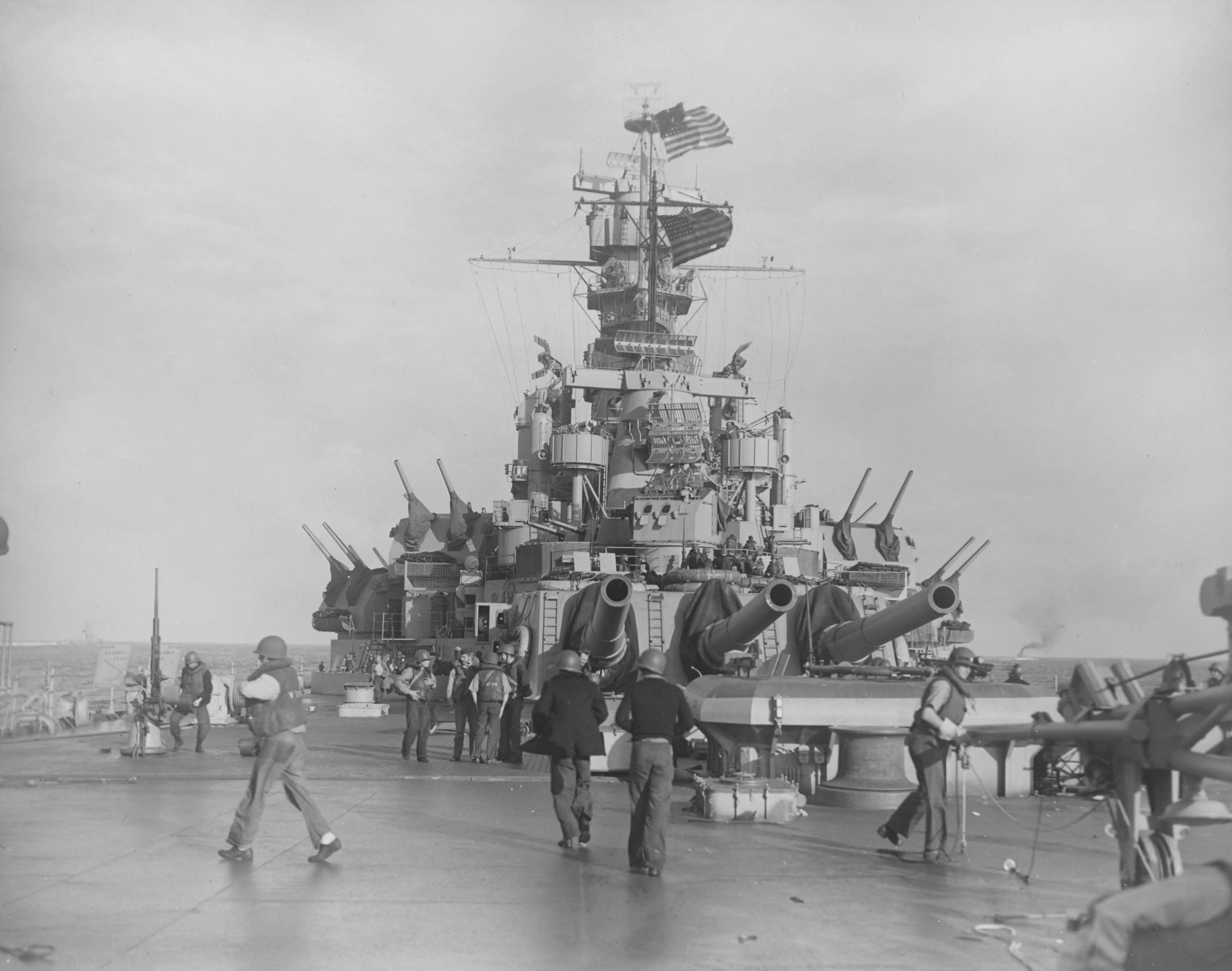 A black and white photograph taken from the stern of USS Massachusetts. Men in life jackets and helmets are moving around the deck, loading guns, and preparing for battle.