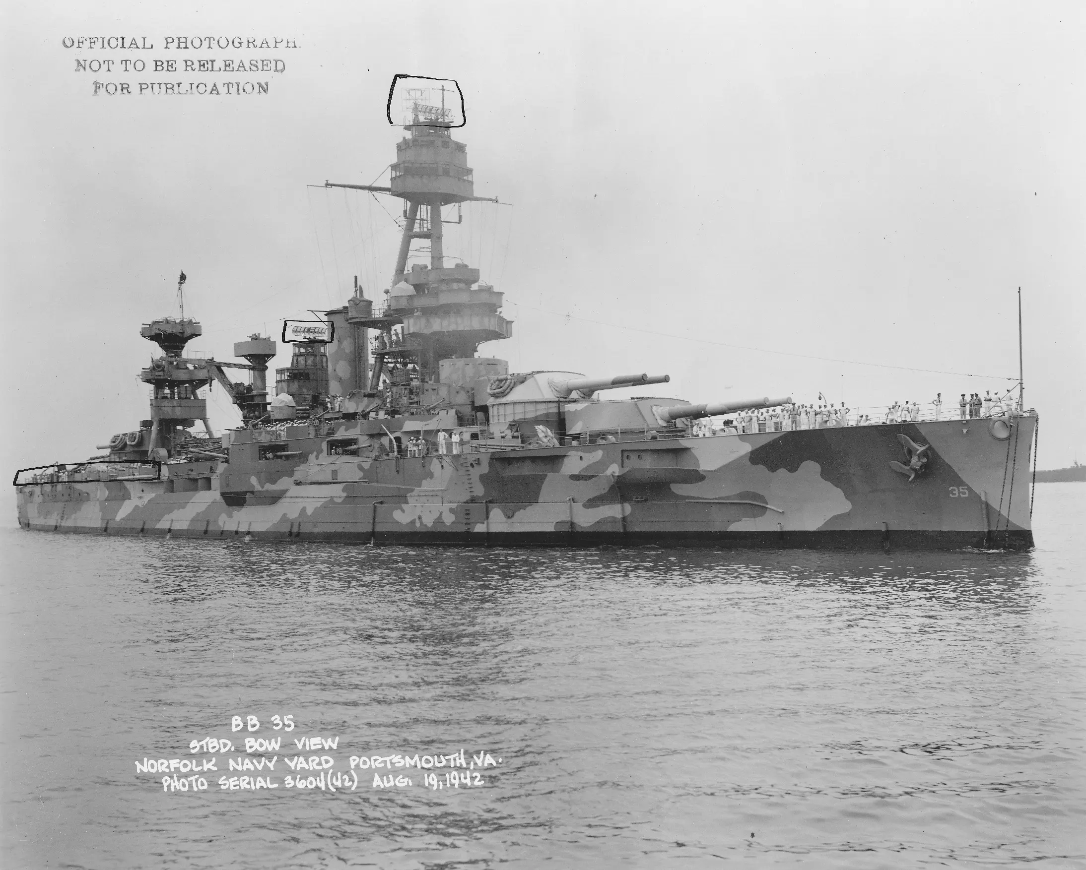 A black and white photograph showing Battleship Texas from the side. The ship is painted in a multicolor camouflage scheme, with loosely vertical splotches of light on dark. Radar antennae are marked by hand with boxes as well as the stern. A stamp in the top left corner reads OFFICIAL PHOTOGRAPH NOT TO BE RELEASED FOR PUBLICATION and a handwritten caption in the bottom left reads BB 35 STBD, BOW VIEW NORFOLK NAVY YARD PORTSMOUTH, VA. PHOTO SERIAL 3604(42) AUG, 19, 1942
