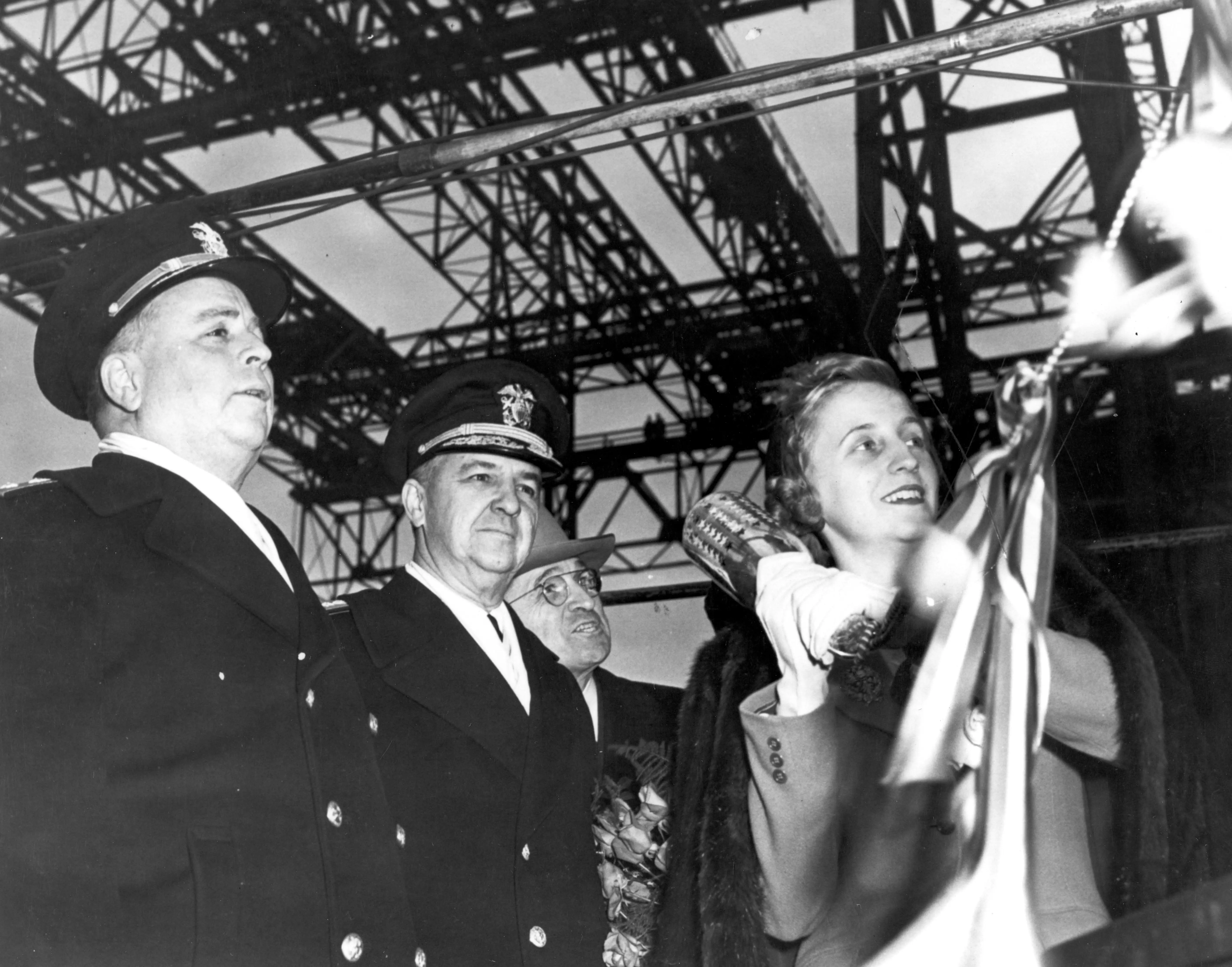 A black and white photograph of a ship launching ceremony, with a woman swinging a champagne bottle as three men watch. Two of the men are admirals in uniform.