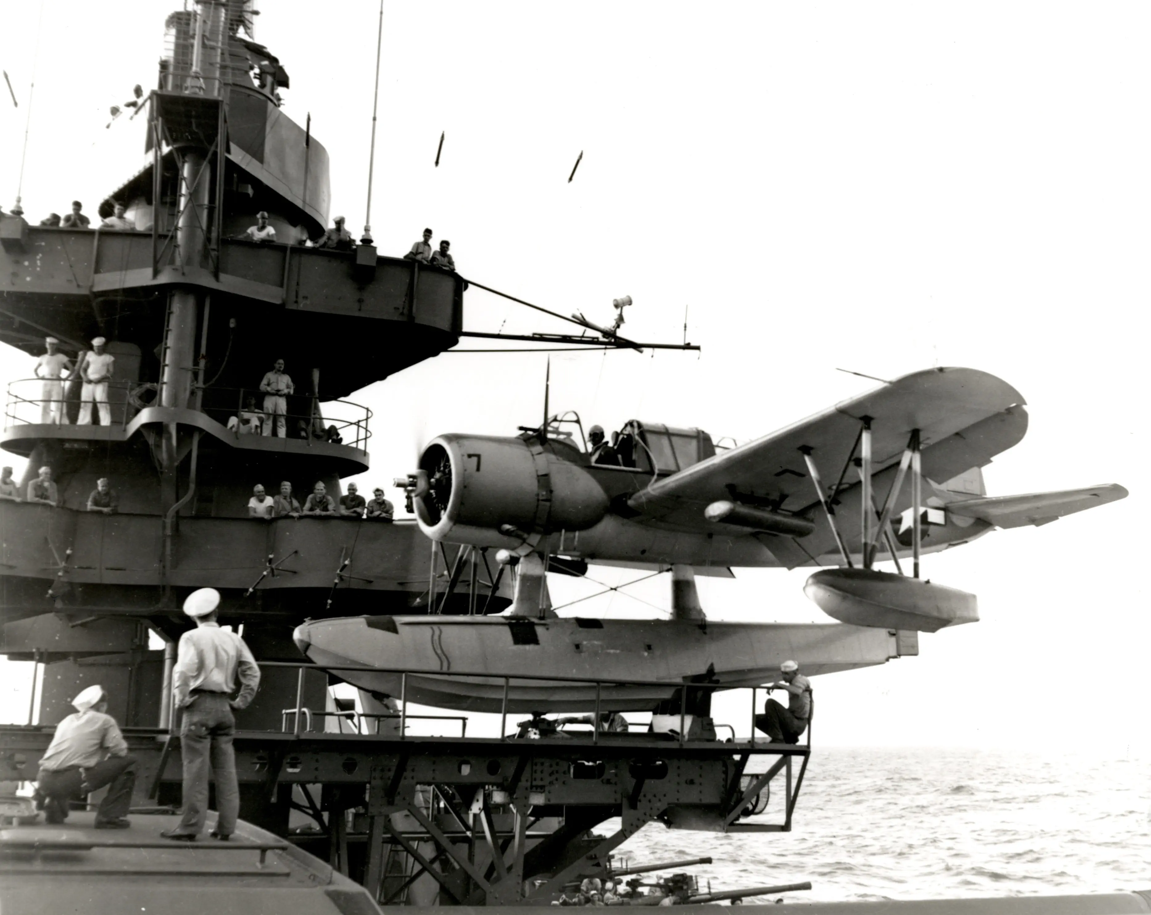 A black and white photograph of a plane sitting on a riveted steel framework. The plane has a large float directly under it, slightly larger than the body of the plane, with two more floats attached to the wings. The framework it rests on is the aircraft catapult on Battleship Texas, with numerous sailors and officers looking on from the main mast and the top of the turret.