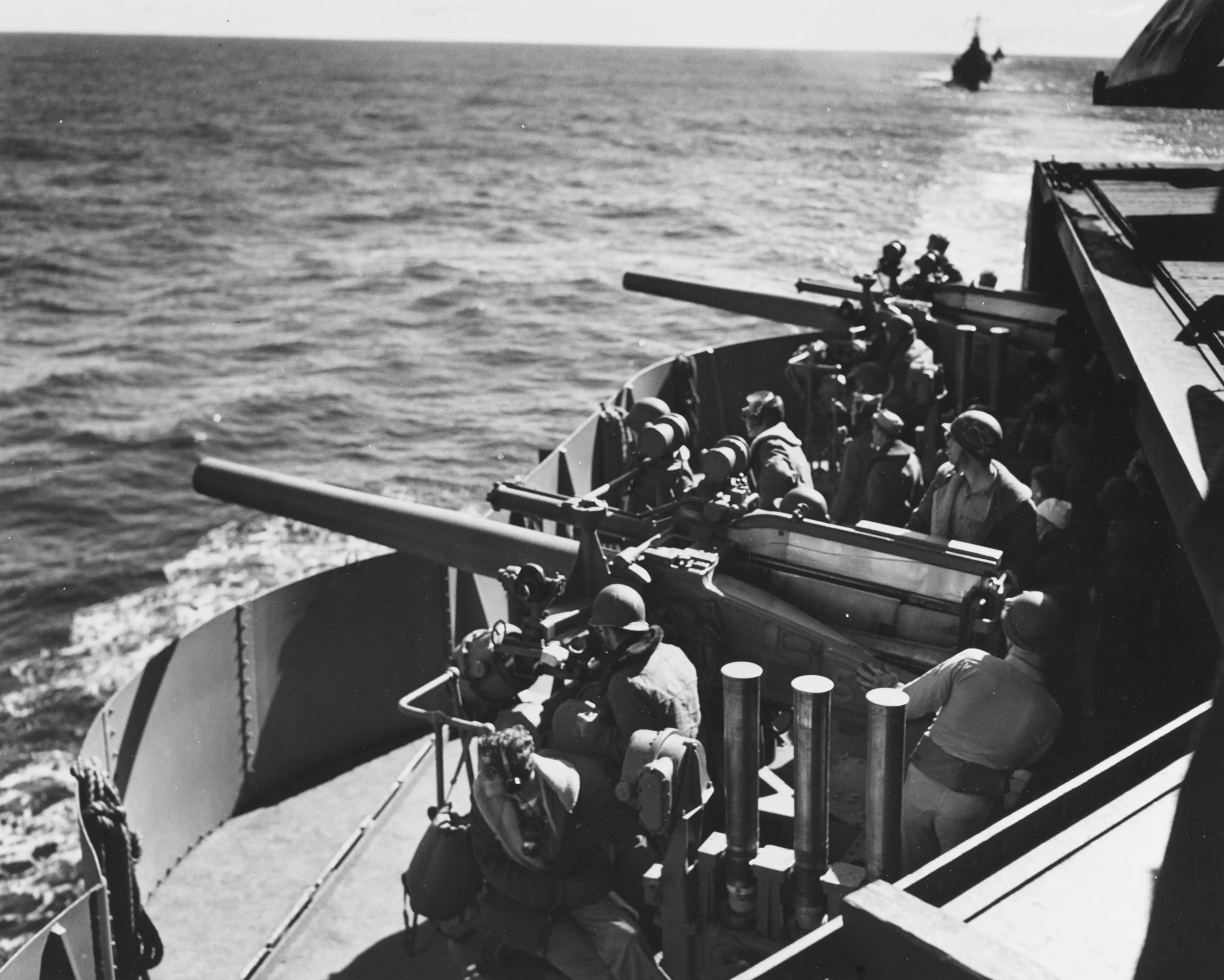 A black and white photograph of American sailors manning two anti-aircraft guns. The guns are trained out to sea with the gun crews mostly looking on and a visible line of other ships trailing behind in the distance. 