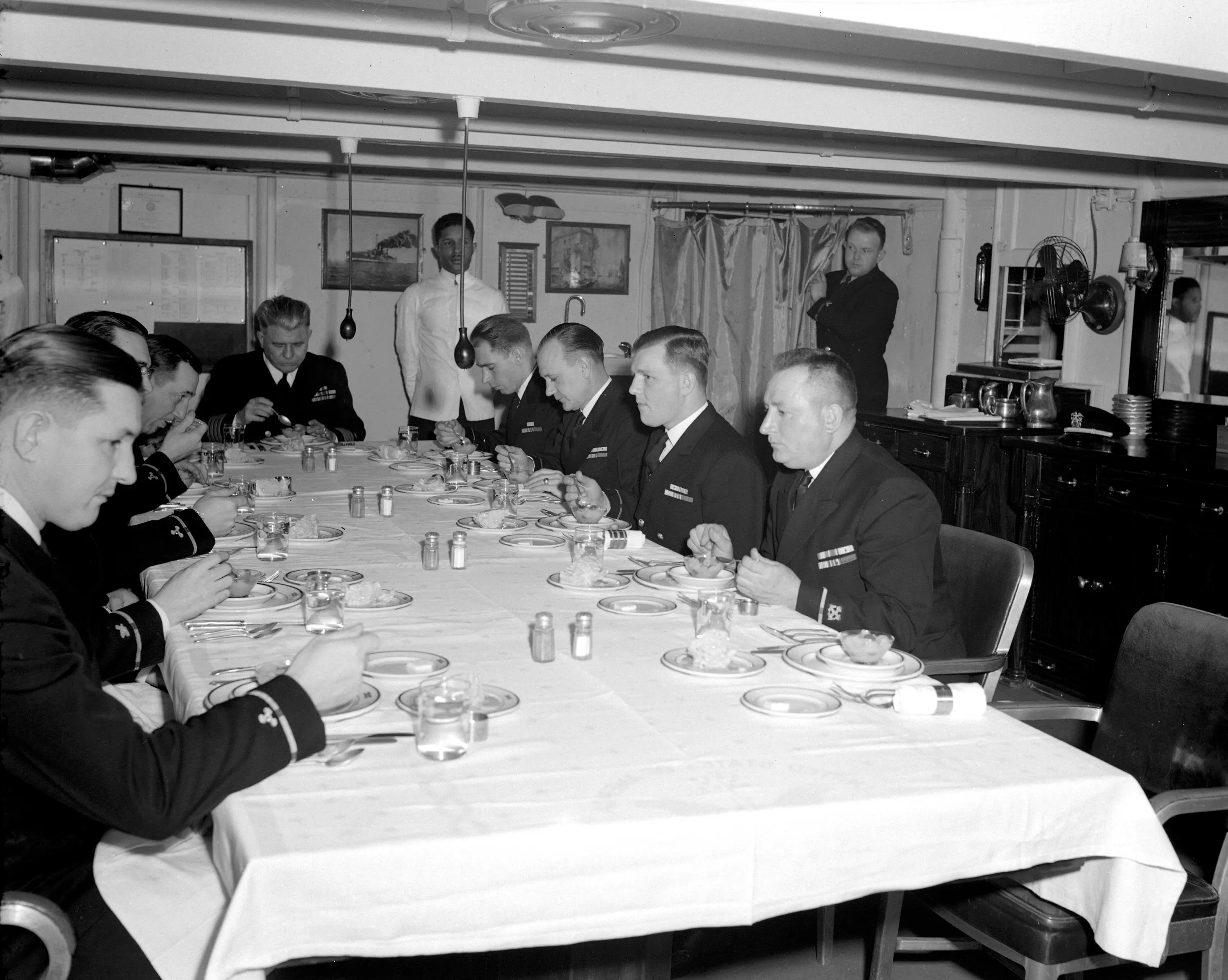 A black and white photograph of warrant officers dining onboard a ship. At the head of the table is the ship's captain. The table is set for formal dining and a Filipino steward in white stands next to the Captain.