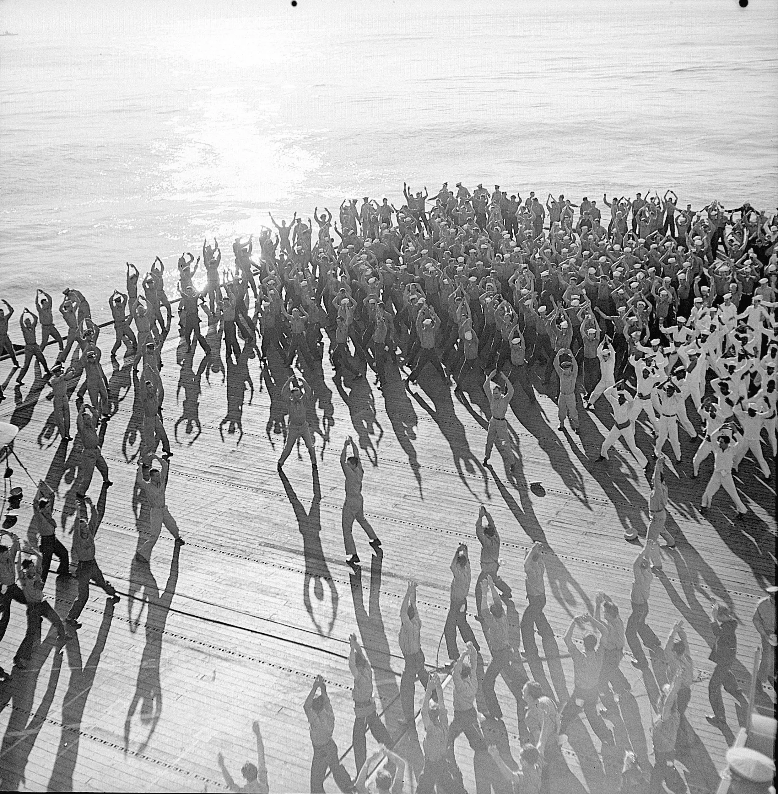 A black and white photograph of the crew of USS Santee doing jumping jacks on the flight deck. Officers in the center appear to be leading the exercises, with over one hundred men around them.