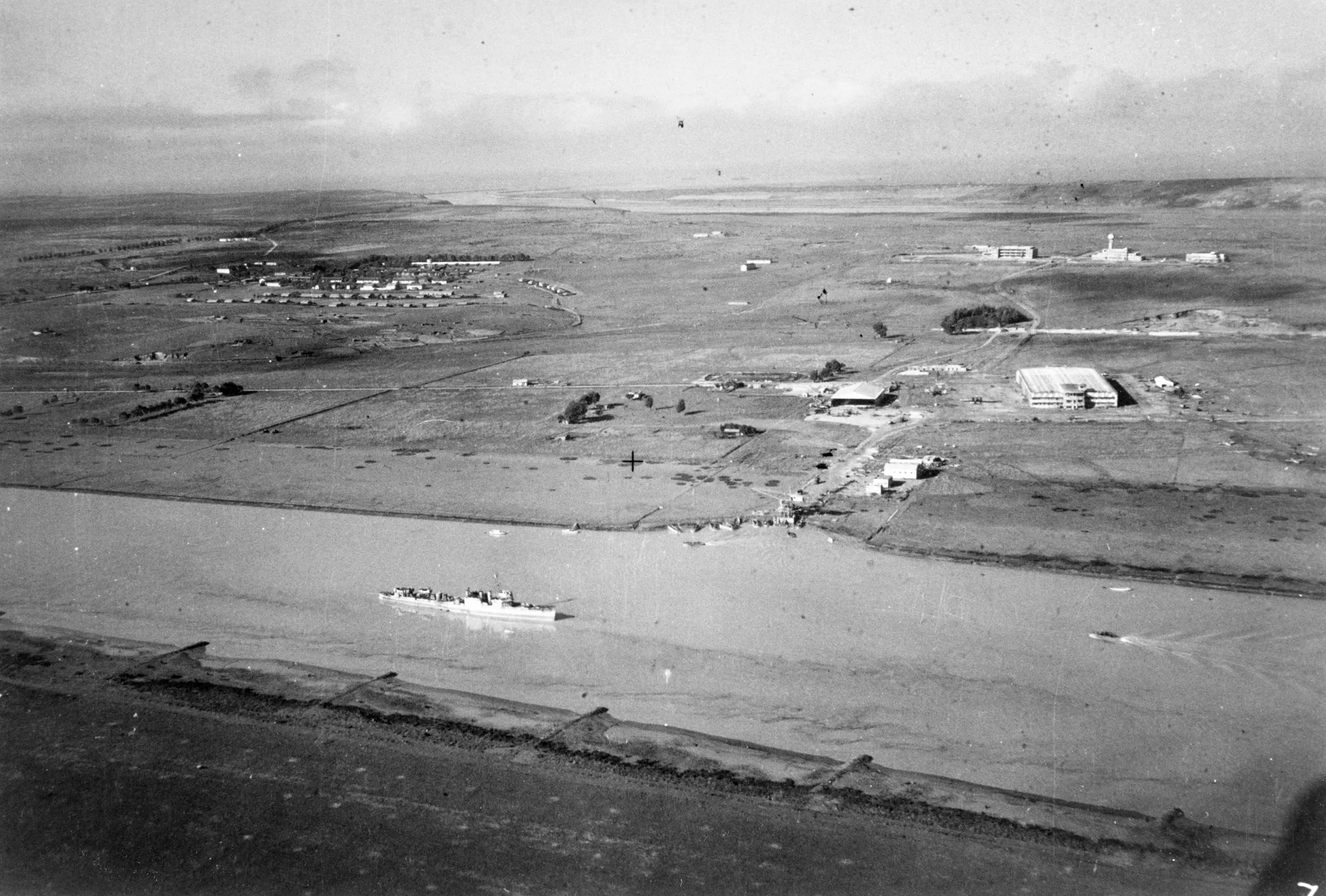 An aerial black and white photograph of a small facility on the banks of a river. In the river is a stationary ship, with several small boats beached on the river bank and a few moving about. The land is mostly flat and empty, with a small number of trees and a few buildings near where the boats landed. In the distance is a larger cluster of buildings and a few more scattered about, but the overall area is empty.